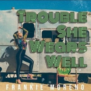 A woman in a colorful top and shiny black pants poses by a vintage trailer with "TROUBLE SHE WEARS WELL" in green.