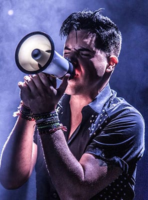 A man with dark hair and a short-sleeved shirt shouts into a white megaphone, his eyes closed.