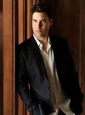 A man with dark hair in a black suit and white shirt leans against a brown wooden wall.
