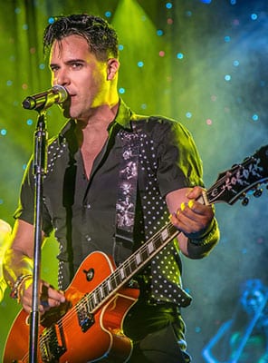 A male musician with dark hair, wearing a black studded shirt, sings into a microphone while playing an orange electric guitar.