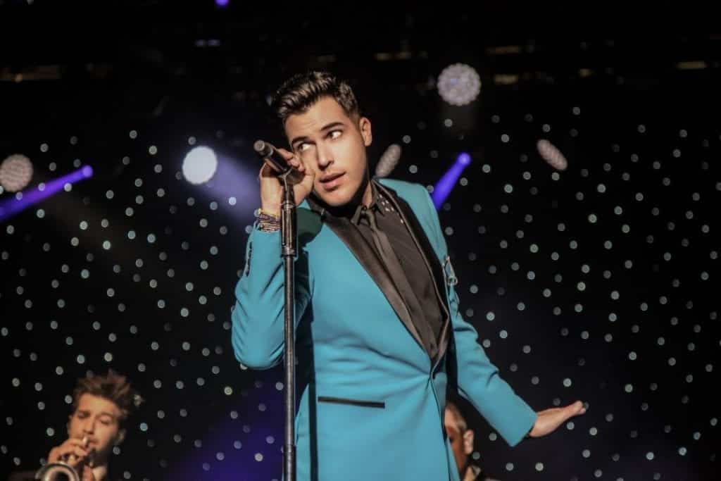 A man in a teal suit with a black lapel holds a microphone, looking up and to the right.