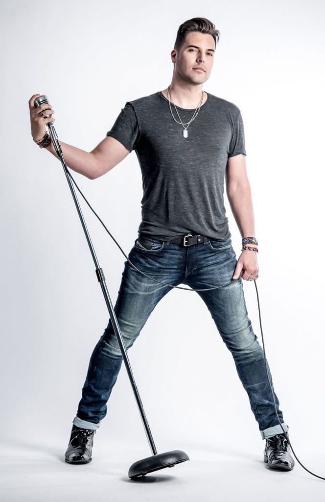 A man in a dark gray t-shirt and jeans holds a vintage microphone on a stand, looking forward.