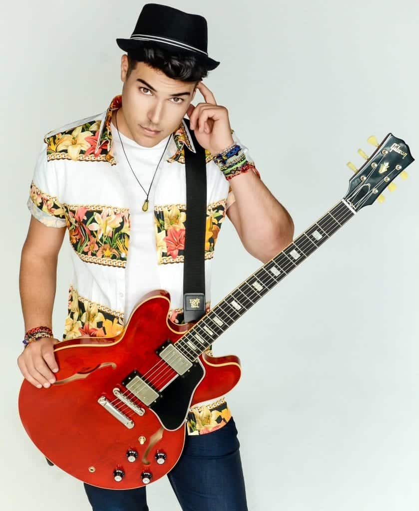 A young man in a black fedora and floral shirt holds a red "Gibson" electric guitar.