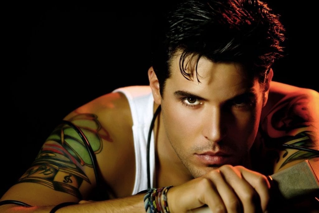 A man with dark hair and brown eyes, wearing a white tank top and colorful bracelets, looks directly at the viewer.