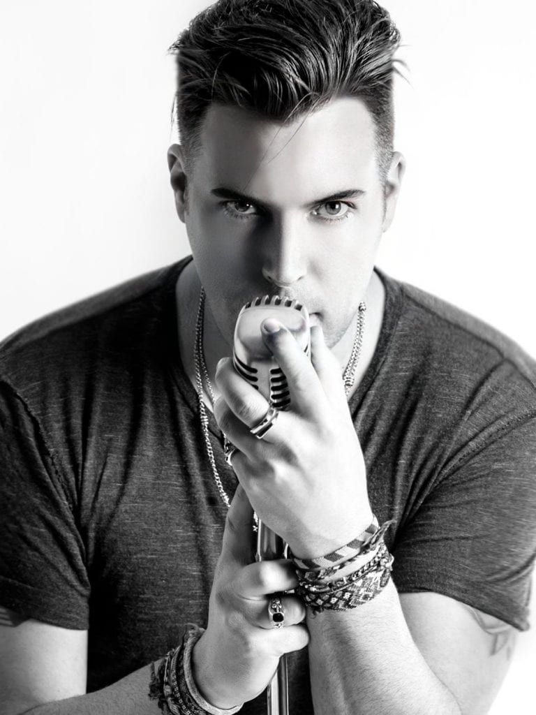 A man in a dark t-shirt and jewelry holds a vintage microphone to his mouth, looking directly forward.