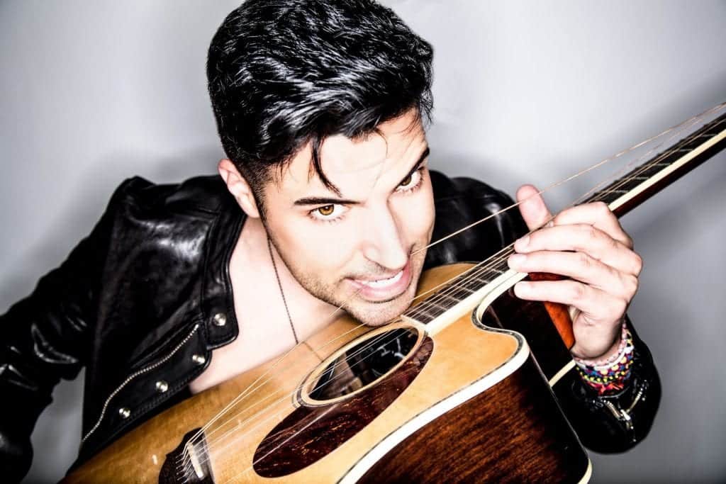 A man in a black leather jacket with dark hair and brown eyes bites a guitar string while playing.