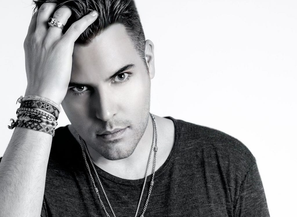 A man in a dark shirt with layered necklaces and bracelets looks intently at the viewer, hand in hair.