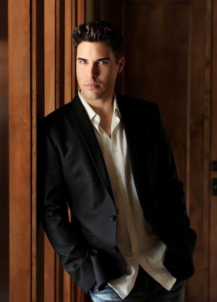 A man with dark hair and bright eyes leans against a wooden wall, wearing a black blazer and white shirt.