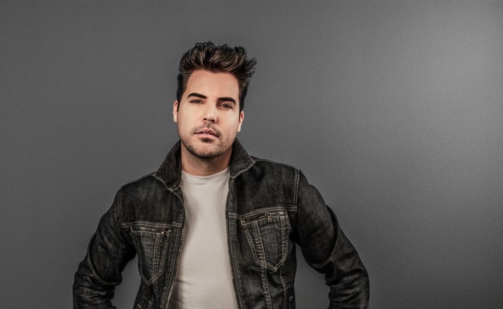 A man with dark hair and a beard, wearing a dark denim jacket over a white t-shirt, looks forward.