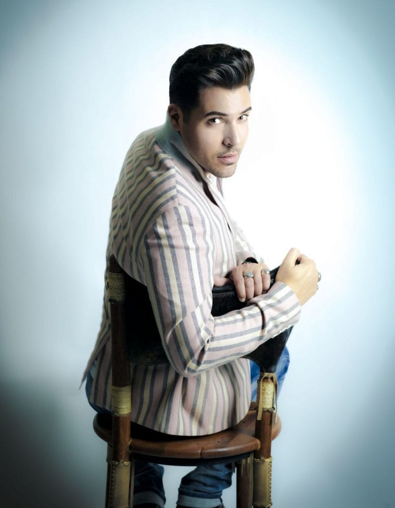 A man with dark hair and a striped pink and gray jacket sits backward on a chair, looking over his shoulder.