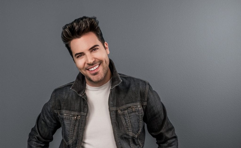 A smiling man with dark hair and a white shirt under a denim jacket poses against a gray wall.