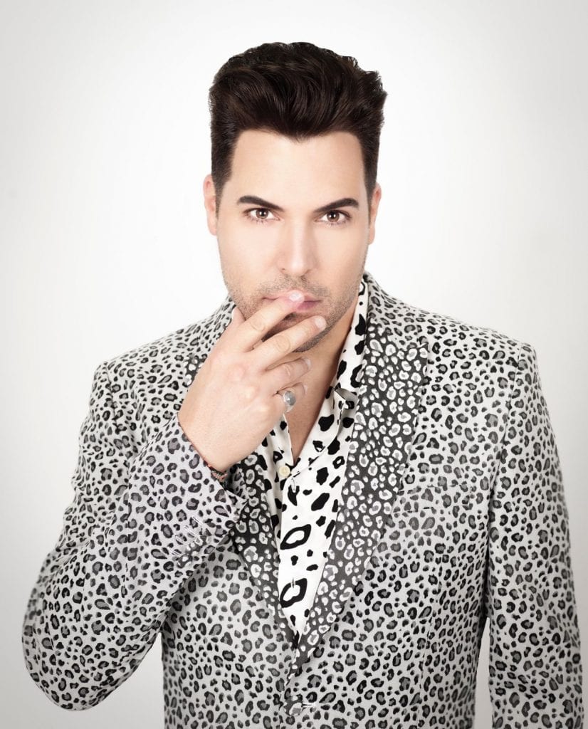 A man with dark hair and a leopard print jacket holds his fingers to his chin, looking forward.