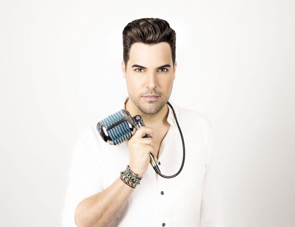 A man with dark hair and a white shirt holds a vintage chrome and blue microphone on his shoulder.