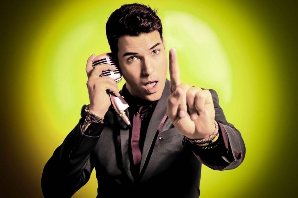 A man in a suit holds a microphone to his ear, pointing his finger up, against a yellow-green background.