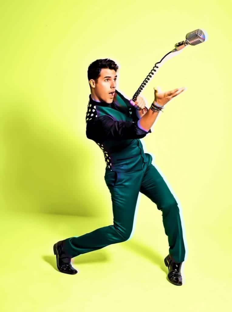 A man in a dark green suit and black shirt with white polka dots holds a vintage microphone.
