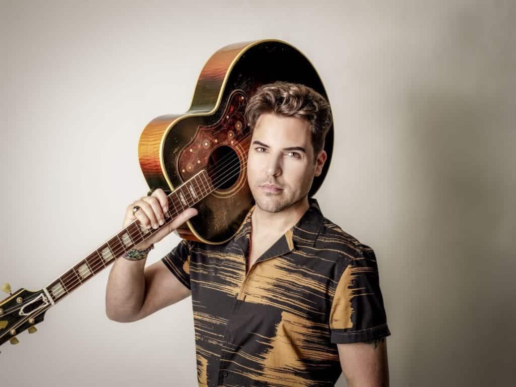 A man with brown hair and a patterned shirt holds an acoustic guitar over his left shoulder.