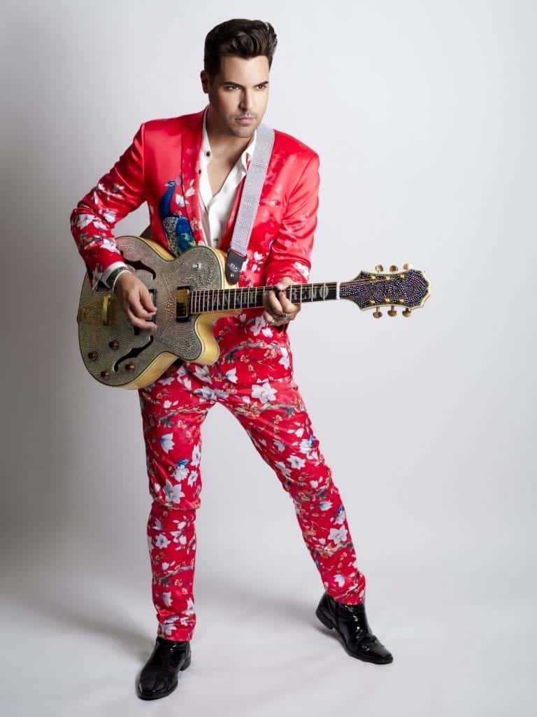 A man in a red floral suit with a peacock design plays a gold electric guitar with a jeweled headstock.