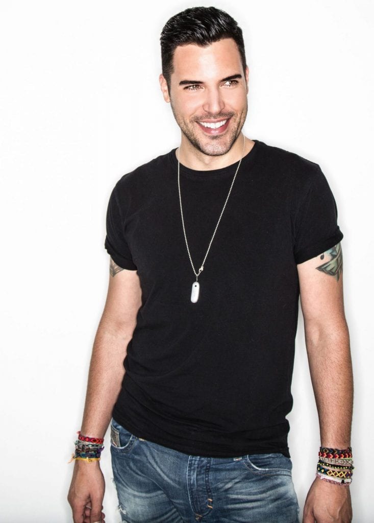 A smiling man with dark hair, a black t-shirt, jeans, and beaded bracelets looks right.