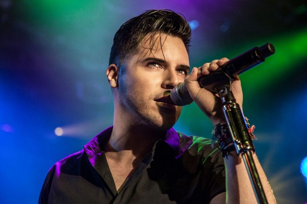 A man with dark hair, wearing a black shirt, sings into a microphone on a colorful stage.