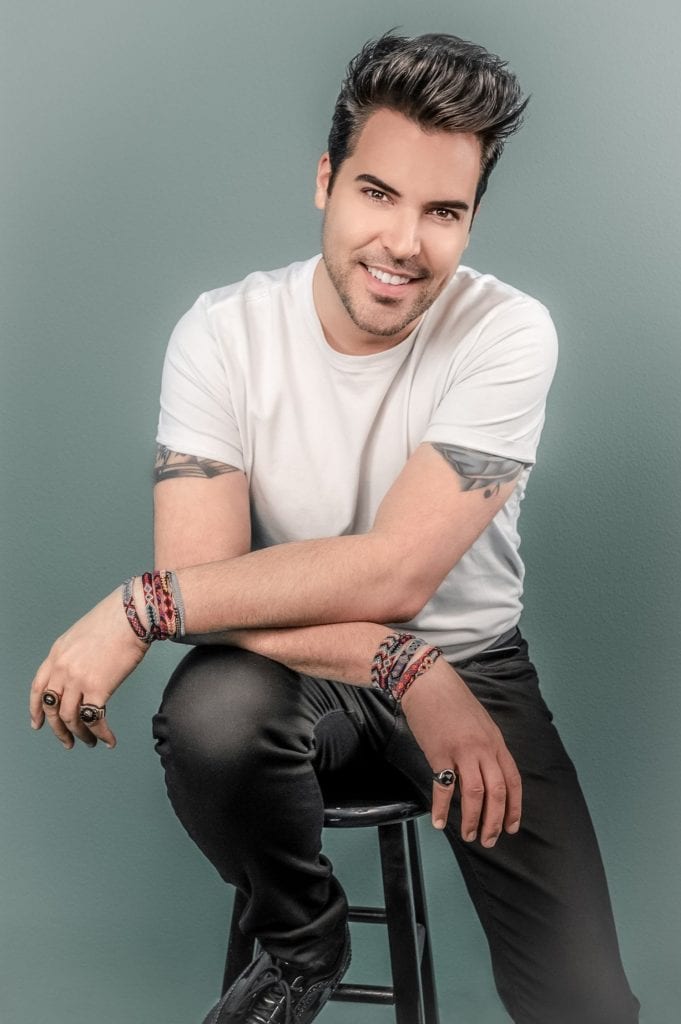 A smiling man with dark hair, a white t-shirt, black pants, and bracelets sits on a stool.