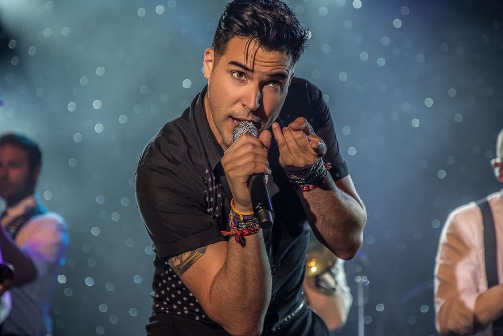A male singer in a black shirt with rolled sleeves holds a microphone and gestures with his left hand.