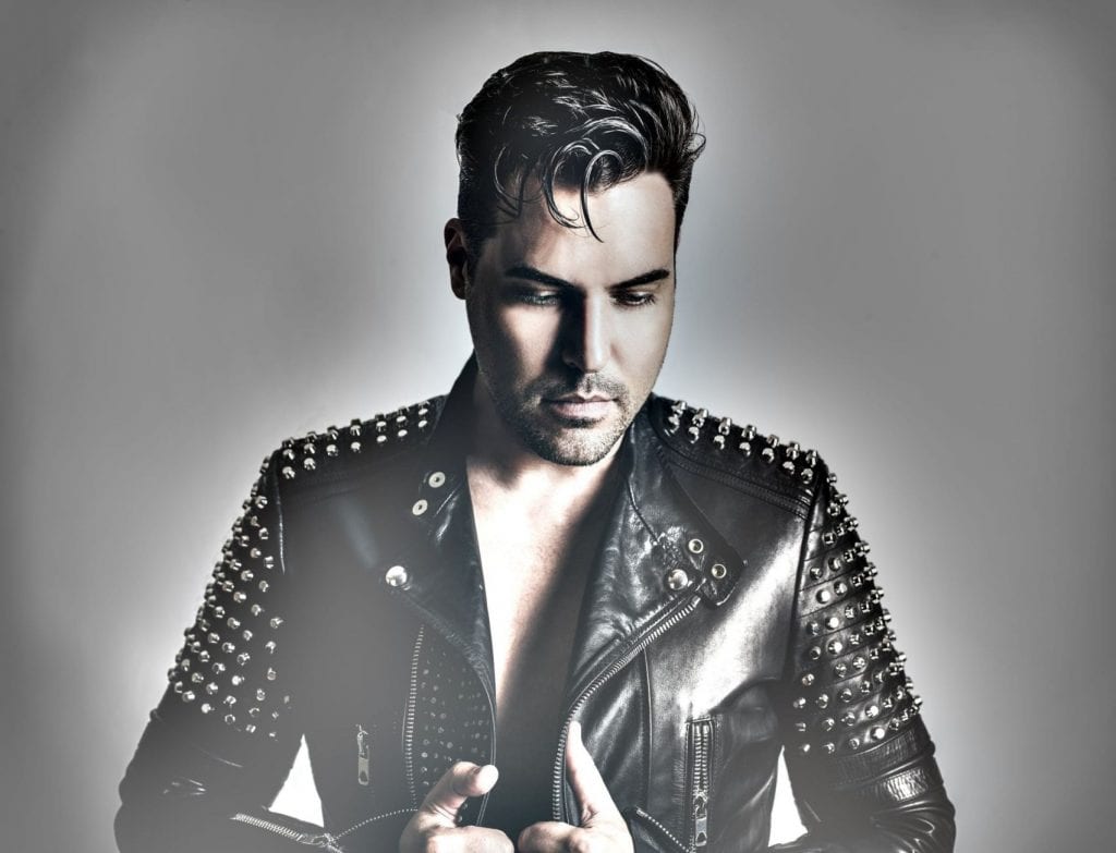 A man with dark hair, wearing a black studded leather jacket, looks down against a gray background.