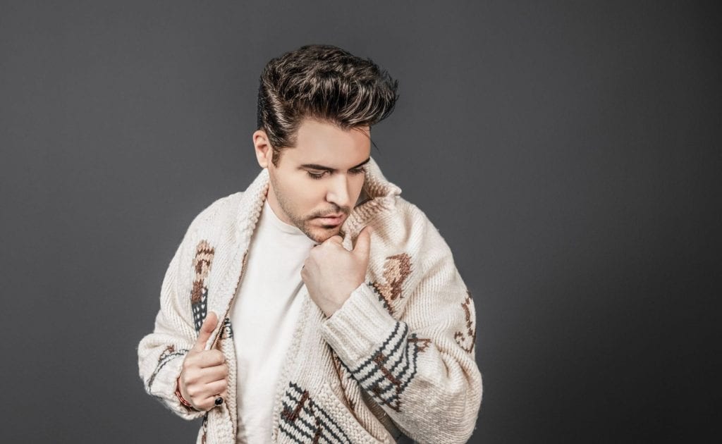 A man with dark hair in a pompadour looks down, adjusting his cream-colored patterned cardigan.