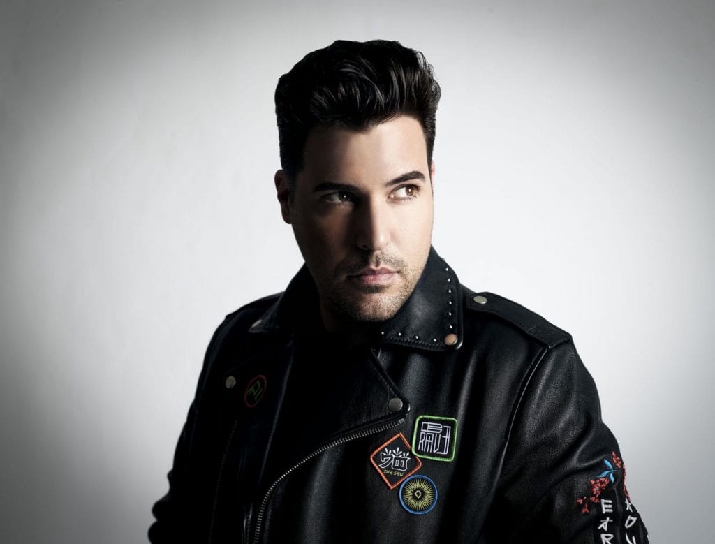 A man with dark hair and a black studded leather jacket with colorful patches looks right.