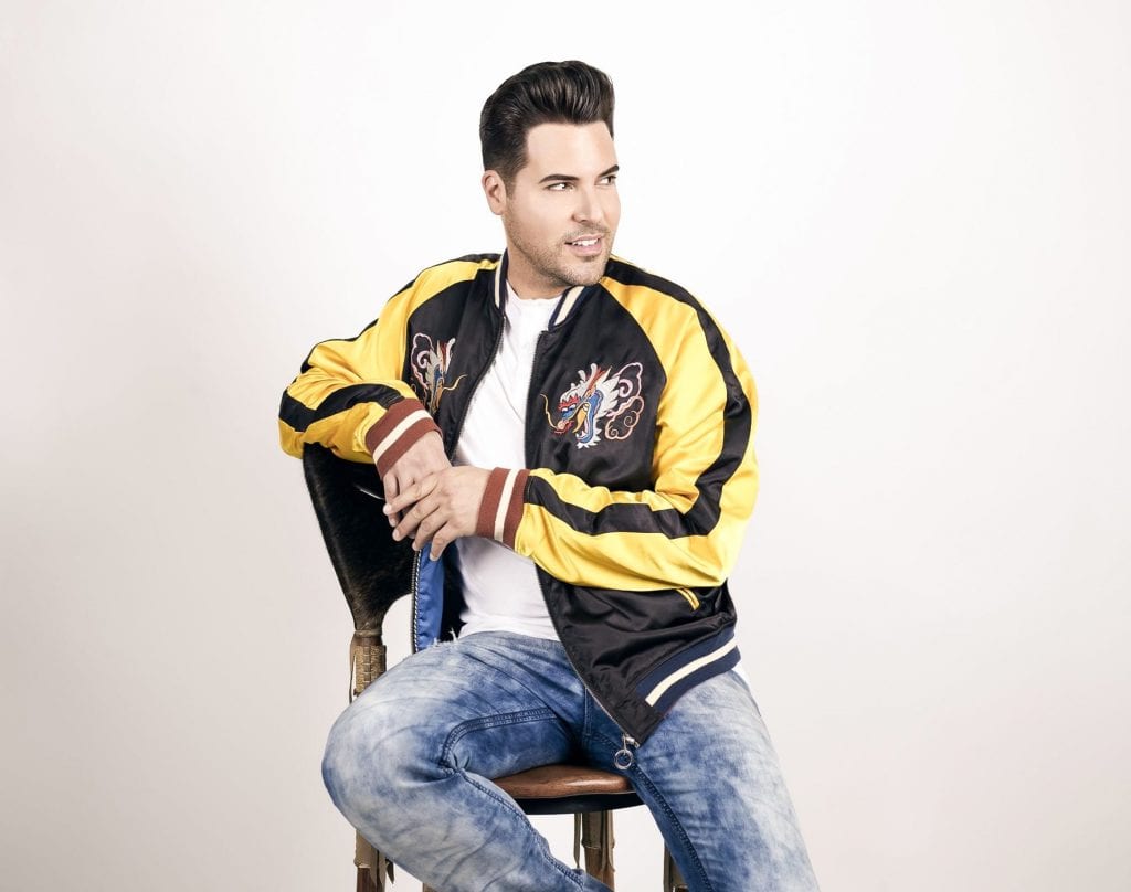 A man with dark hair, wearing a black and yellow jacket with embroidered dragons, sits on a chair.