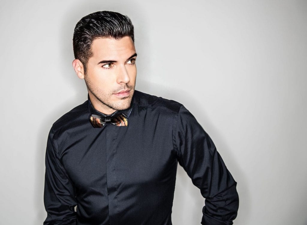 A man with dark hair and a patterned bow tie wears a black shirt, looking right against a white background.