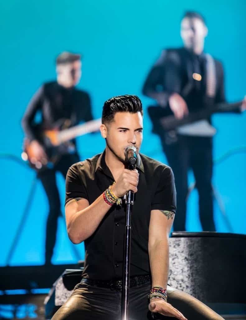 A male singer in a black shirt holds a microphone, with two blurred guitarists behind him.