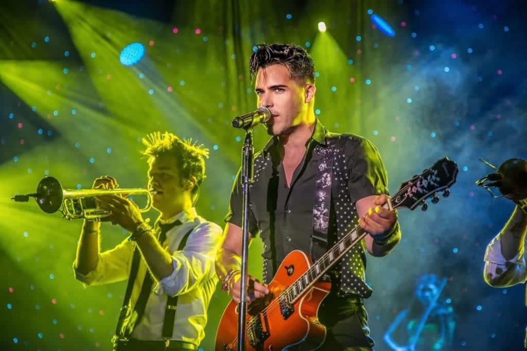 A male singer with a guitar and a trumpeter perform on stage under green and blue spotlights.
