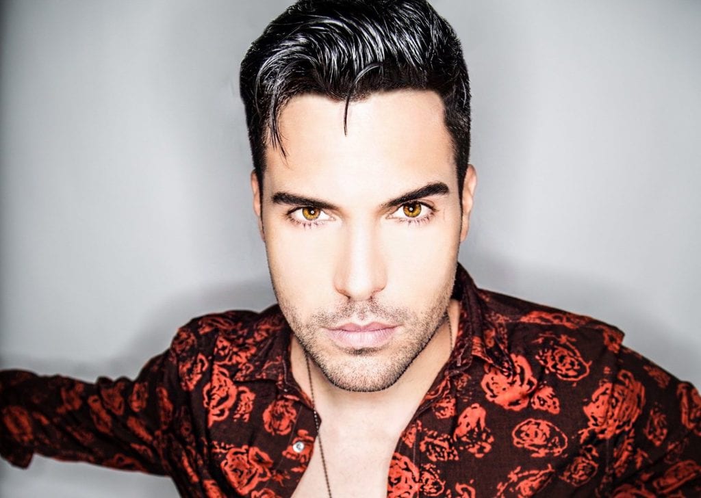 A man with dark hair, light skin, and amber eyes stares forward, wearing a dark red floral shirt.