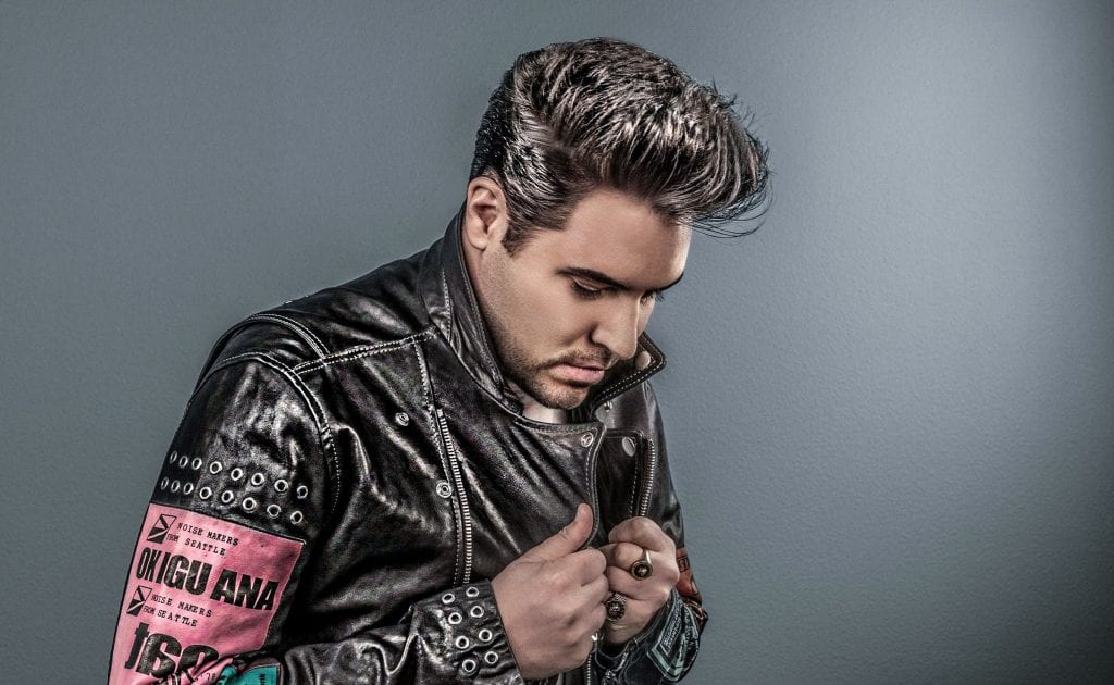 A man with a pompadour hairstyle and a black leather jacket with "OKIGU ANA" on a pink patch.