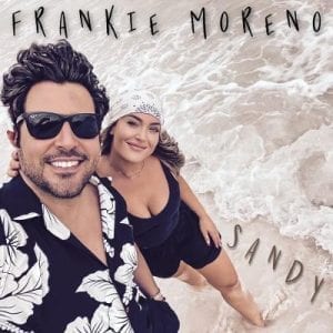 A smiling man in sunglasses and a woman in a bandana stand in shallow water with text "FRANKIE MORENO SANDY".