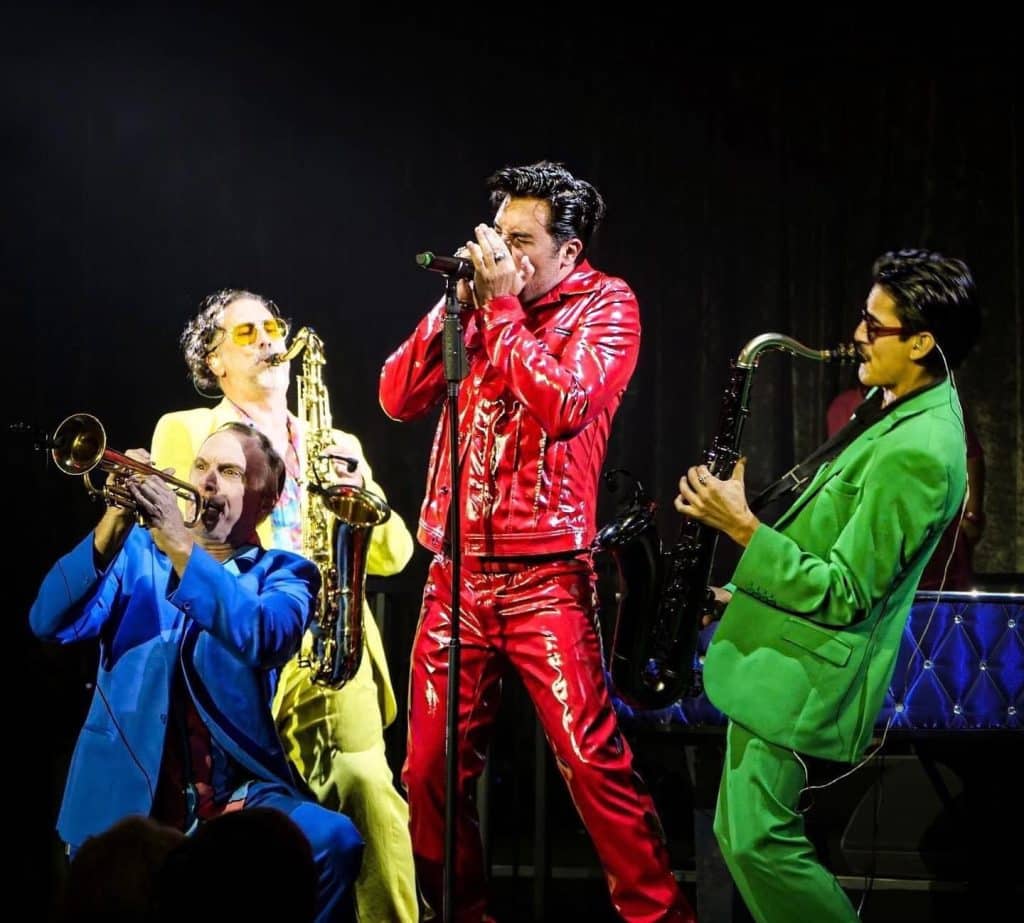 A band performs on stage: a singer in red, a saxophonist in yellow, a trumpeter in blue, and another saxophonist in green.
