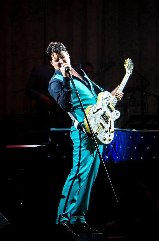 A male singer in a teal suit and black shirt holds a sparkly white guitar and sings into a microphone.