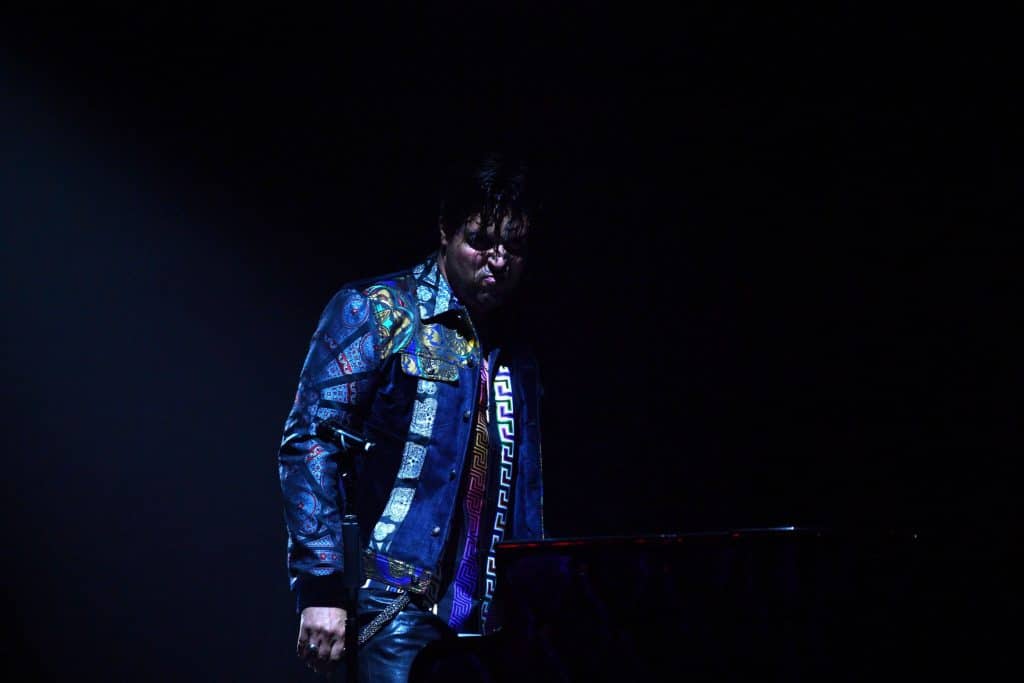 A man in a patterned blue jacket and black leather pants stands in dim light, looking down at a piano.