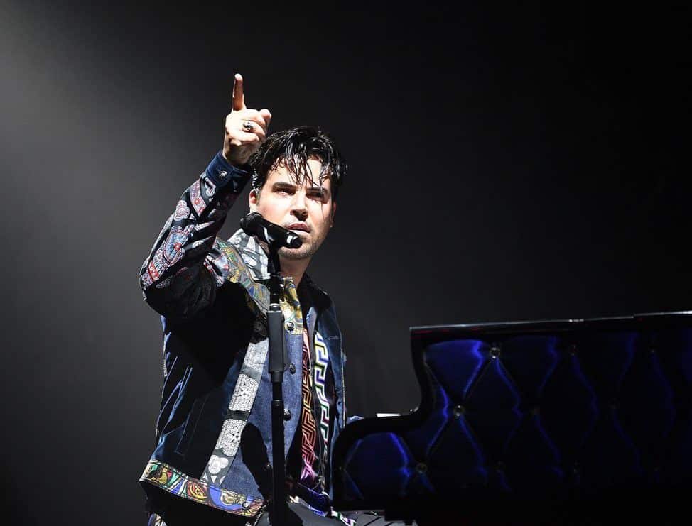 A man with dark hair in a patterned jacket points his finger up while sitting at a dark blue piano.
