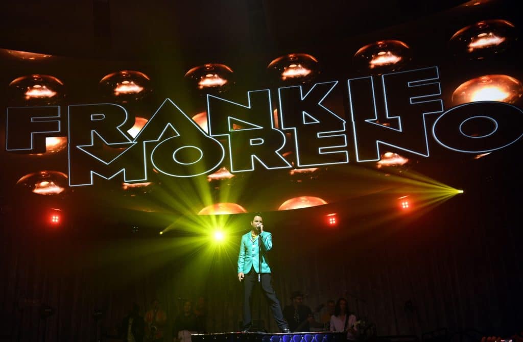 A man in a teal jacket sings into a microphone on stage under bright yellow lights and "FRANKIE MORENO" on a screen.