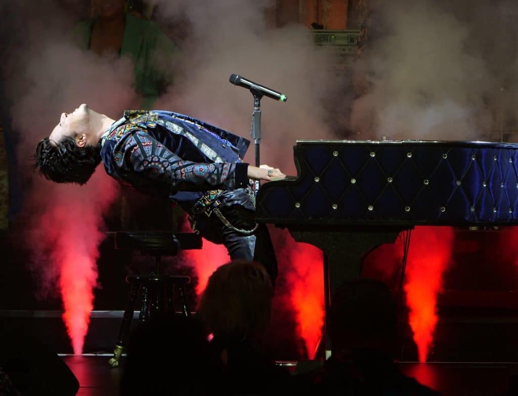 A performer in a patterned jacket leans back dramatically while playing a dark blue, studded piano on a smoky stage with red lights.