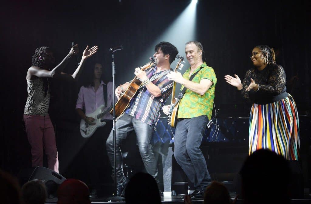 A band of five musicians performs on a dark stage, two guitarists center, with a spotlight overhead.