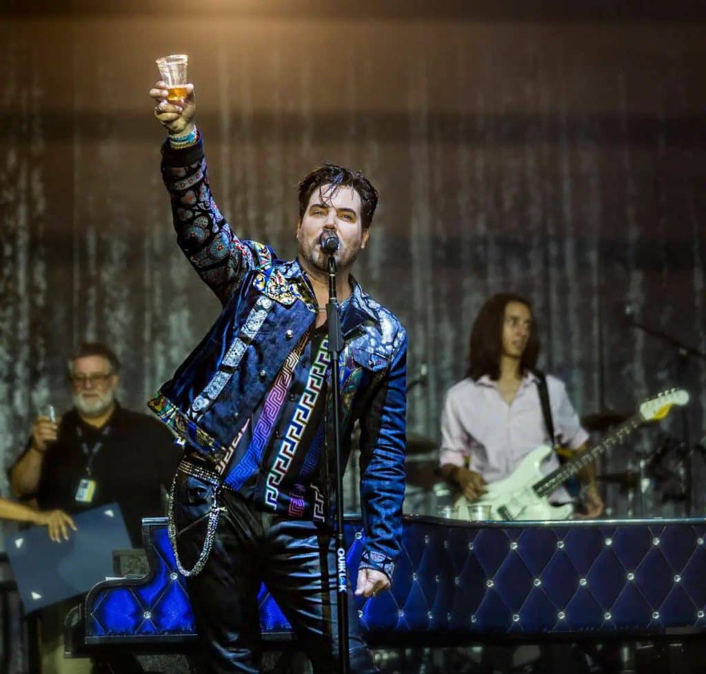 A male singer in a blue patterned jacket holds a drink aloft, with musicians and a piano behind him.