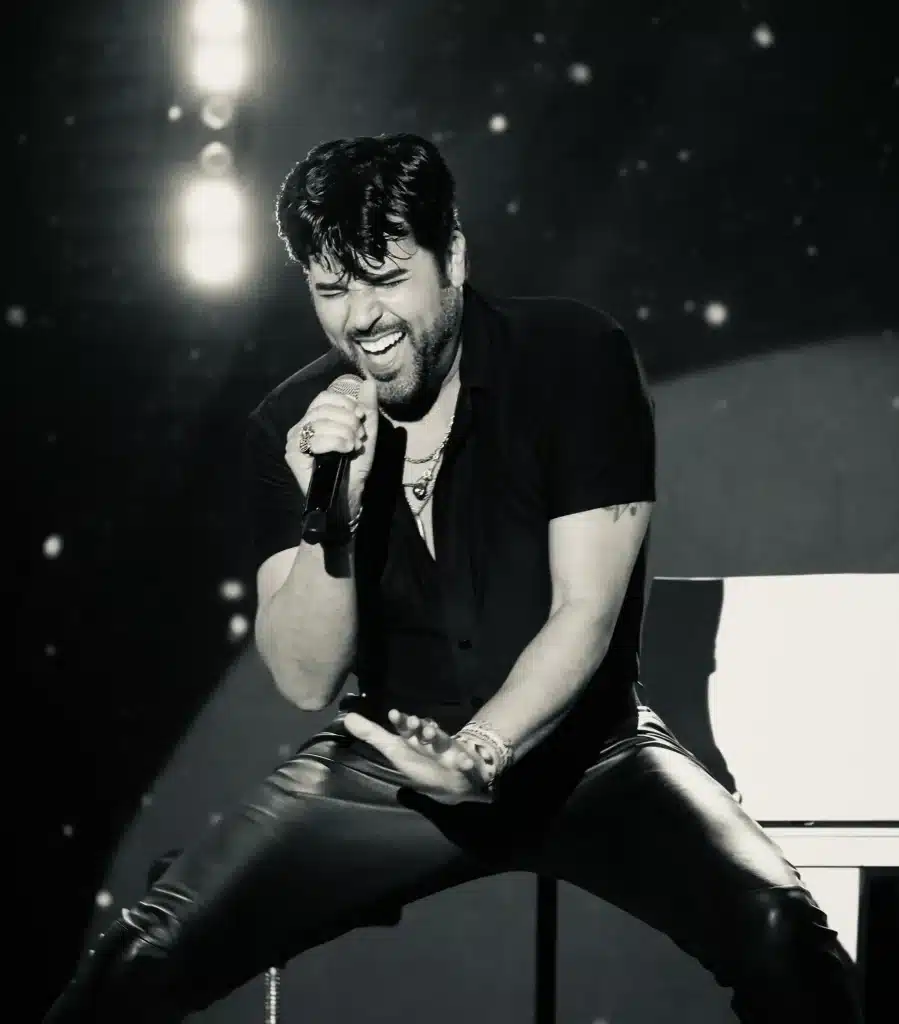 A man with dark hair and a beard, wearing a black shirt and leather pants, sings passionately into a microphone.