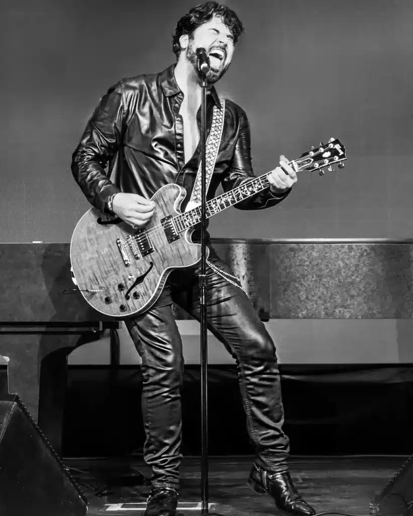 A man in a leather jacket and pants sings into a microphone while playing an electric guitar.