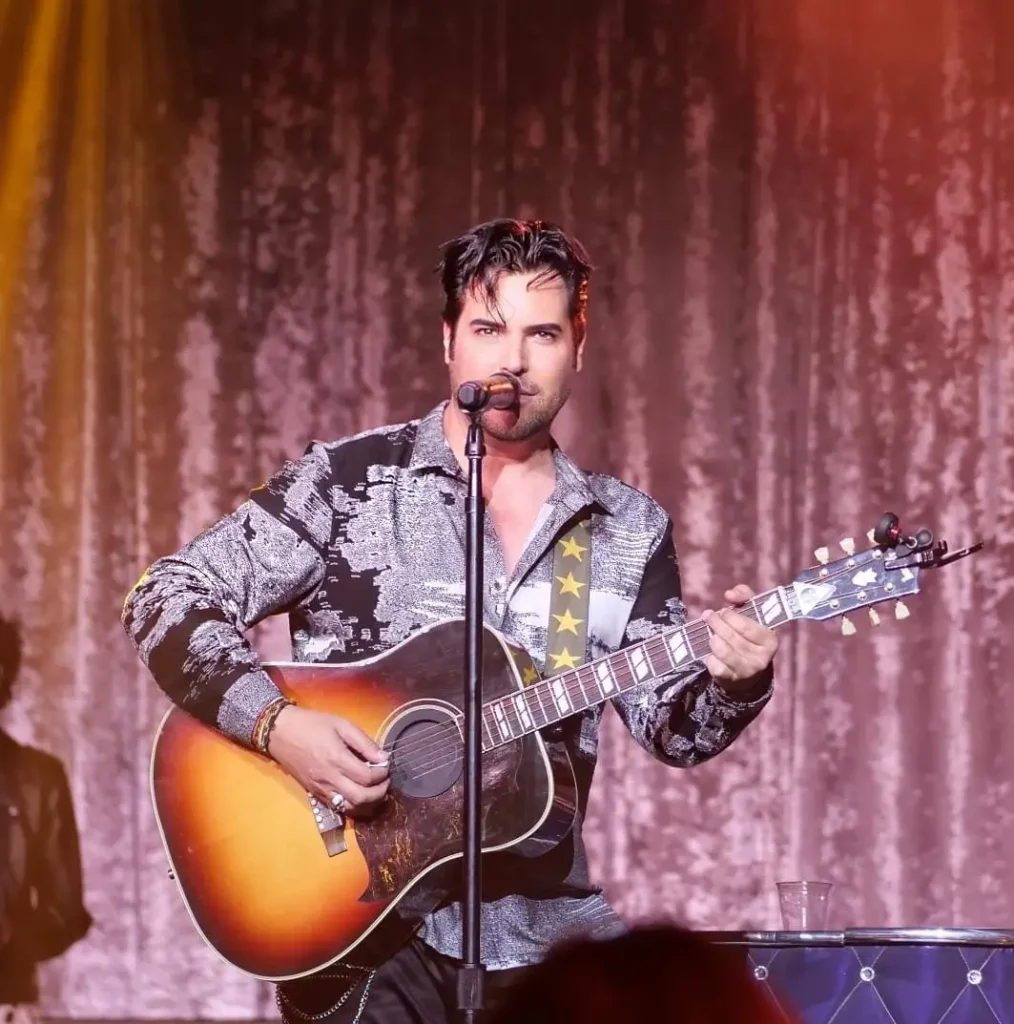 A man with dark hair and a patterned shirt sings into a mic while playing an acoustic guitar.