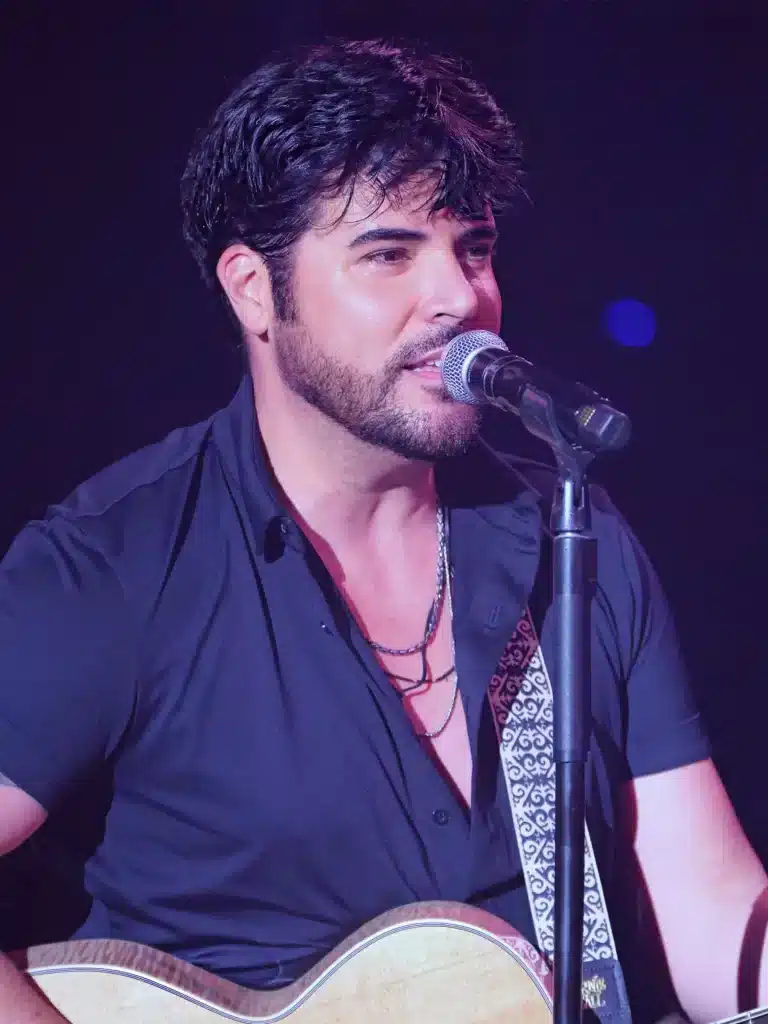 A man with dark hair and a beard sings into a microphone while playing an acoustic guitar.