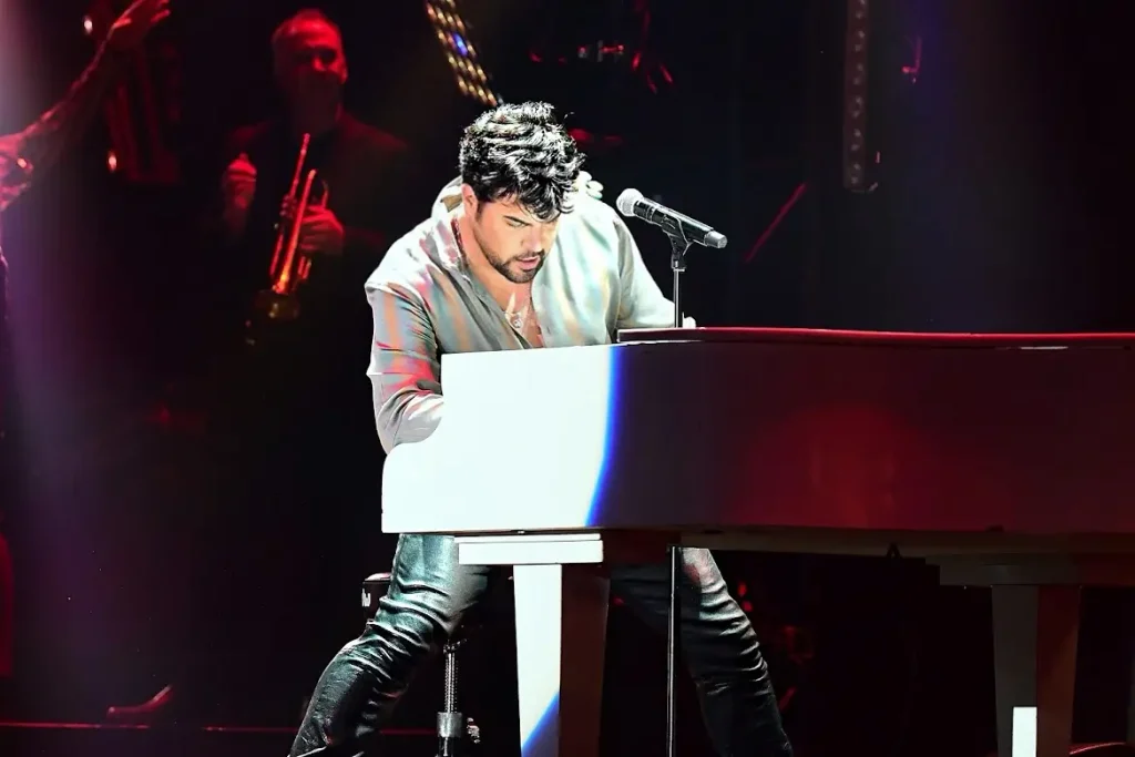 A man with dark, wavy hair plays a white piano on stage under blue and red lights, with a trumpeter in the background.