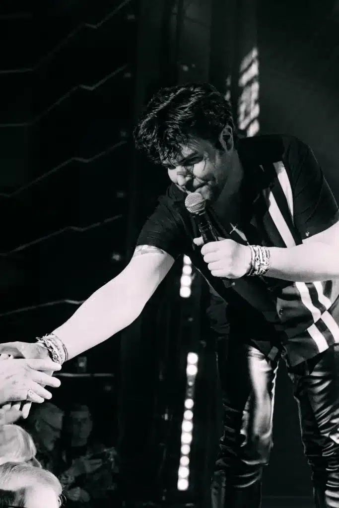 A man in a striped shirt and leather pants holds a microphone while shaking a fan's hand.