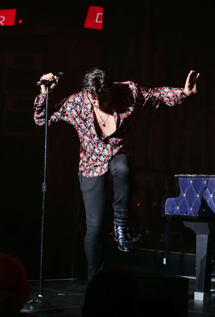 A male singer in a patterned shirt, black pants, and shiny shoes holds a microphone, mid-jump on stage.